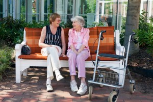 Two elderly female friends sitting chatting on a park bench under the shade of a tree one using a walking aid.